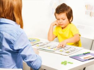 Child and adult sitting at a table. The kid has their head on their hand and is pointing to a box on a sheet of paper.