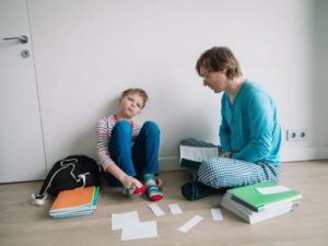 Adult and child sitting on the floor with a multiple stacks of books nearby. The child is staring forward, and the adult is looking at the child while holding an open book.