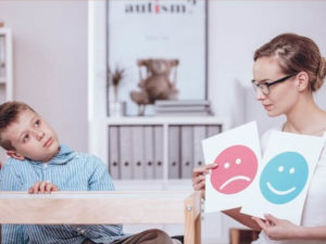 An adult and a child sitting near a wooden table. The adult is holding two cards, one with a smile and one with a frown. The child is staring off to the side.