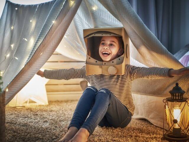Child sitting under a grey blanket tent with fairy lights on it. They are wearing a cardboard cut-out of an astronaut helmet.