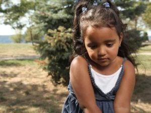 Close-up on a child with ponytails, staring towards the ground. They are standing in front of a pine bush.