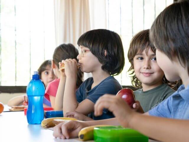 Boy in a school cafeteria, sitting in a line of other kids, looking at the sandwich he is eating.