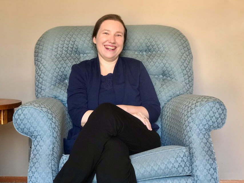 A pale skinned woman with brown hair and blue eyes, smiling at the camera while sitting on a blue textured sofa.