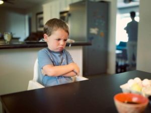 A child sitting in a high chair, crossing their arms and frowning.