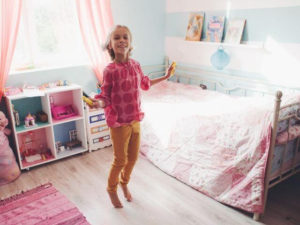 Child, with long blond hair, jumping in a pink and blue themed bedroom.