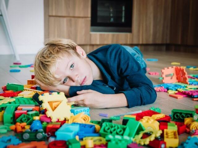 Child laying on the floor in front of a load of Lego pieces scattered about. The child is staring at the camera.