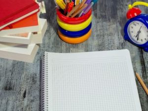 Close-up on a notebook, with a pencil to the side. There's an alarm clock and a cup of pencils above it.