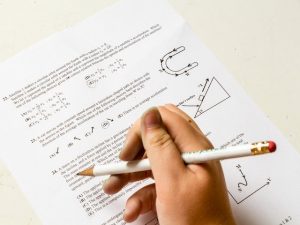 Close-up on a hand holding a pencil above paper with math questions on it.