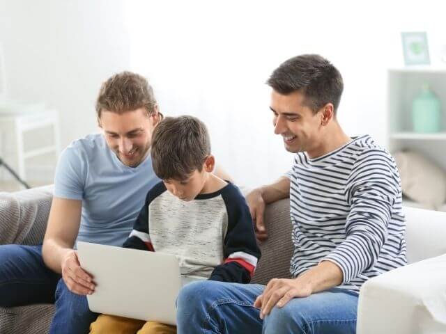 Two adults and a child sitting on a white couch, all looking at a silver computer the child has in their lap. The adults are smiling.