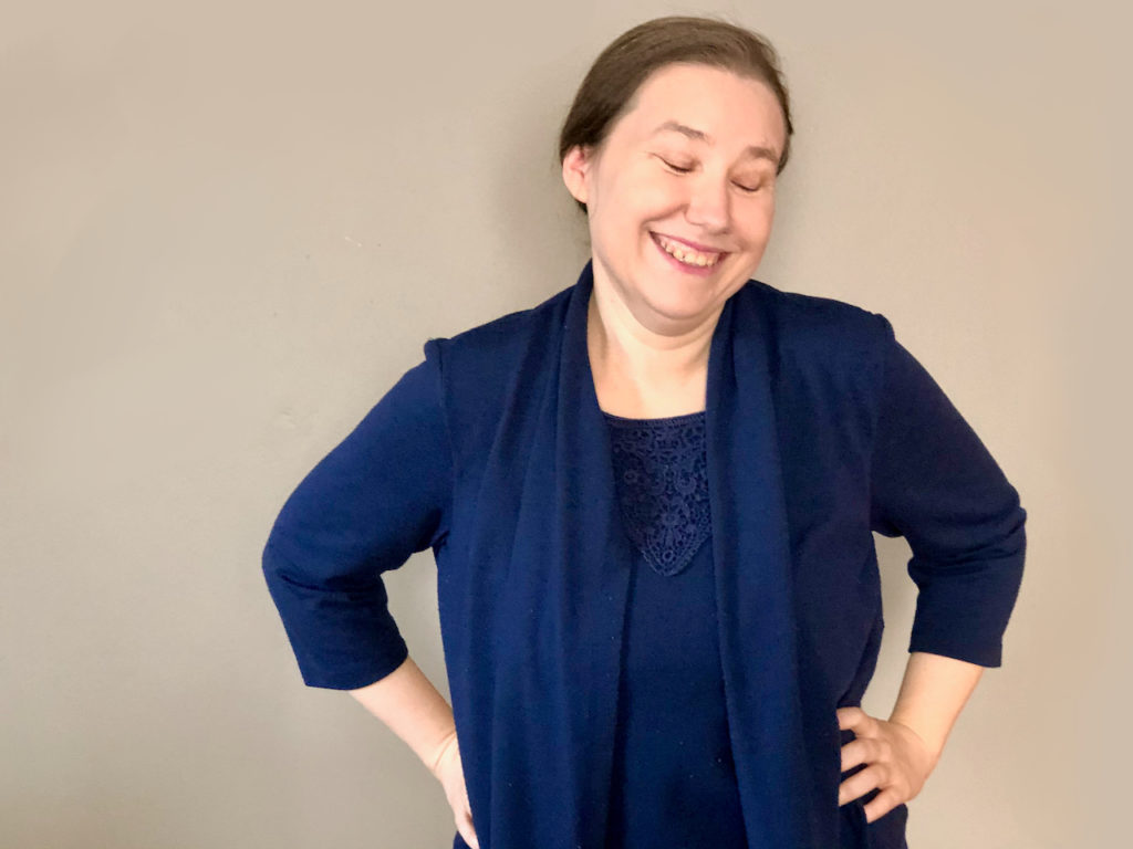 Women in a blue shirt, holding her hands on her hips. Smiling with her head tilted to the side.