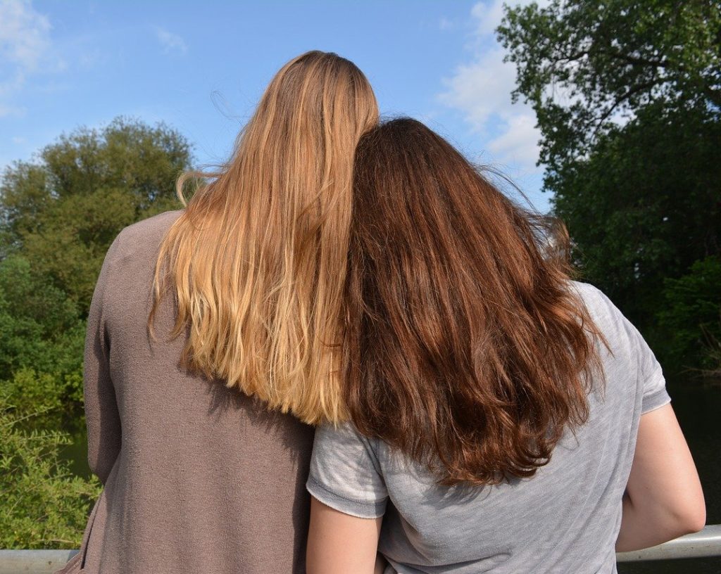The backs of two people with long hair, leaning up against each other, in front of a forest.