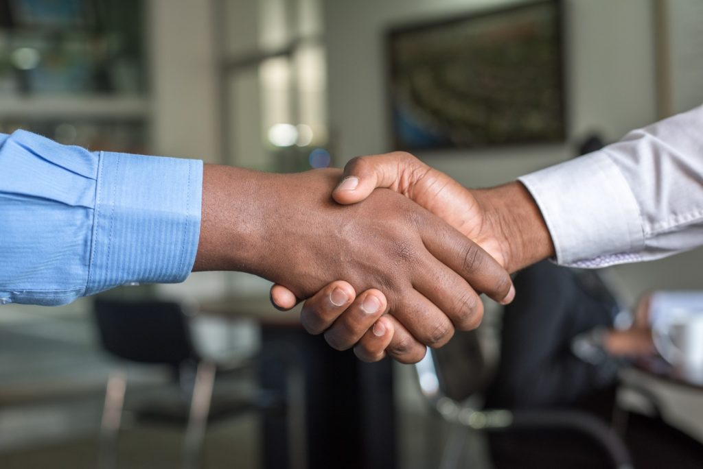 Closeup of the arms and hands of two people shaking hands. Both are wearing business shirts over dark brown skin.