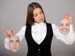 A young woman in a white shirt and black vest holds up a life-like face in each hand as if it were a mask. Each face wears a different expression. She looks to one as if she is deciding between the two.