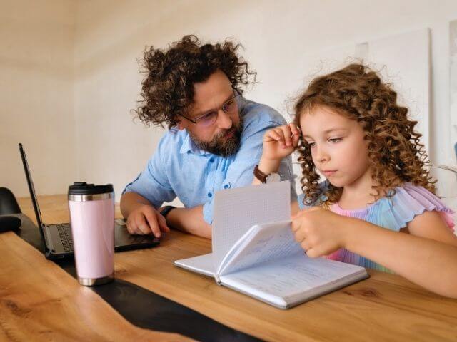 Kid with long curly brown hair at a table, flipping through a book. There is an adult in front of a computer to the kid's side, and they are looking at the kid.