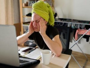 Person with pale skin and a green towel wrapped around their hair is pressing up against their face with their hands. Their eyes are closed and are facing towards a computer at a desk.