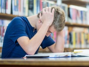 Person with pale skin and blond short hair has their hands on their head. They are facing down towards a table that has a book open on it. There is a bookshelf in the background.