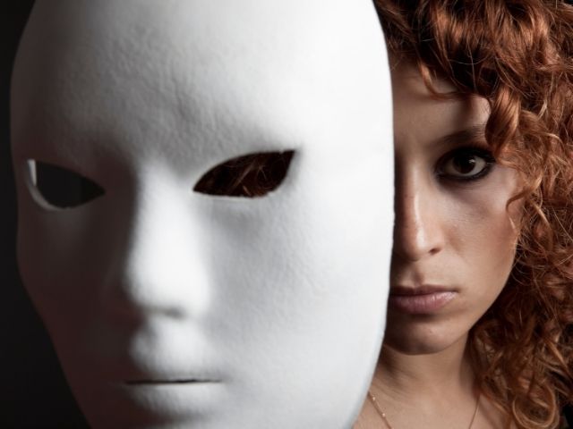 Face of a woman with pale skin and red curly hair. In front of her and hiding half of her face is a white theater mask.