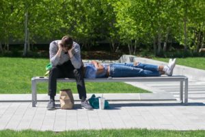 Two people on a bench with bushes behind them. One person is laying down with their head up against the other person, who is sitting and looking into a brown bag.