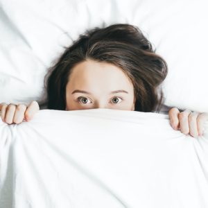 Woman in bed with sheets pulled up over her face so just her eyes and forehead are visible. Her pale skin and dark brown hair contrast with the bright white of the sheets and pillow.