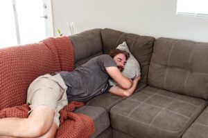 Man wearing a t-shirt and shorts, with pale skin and dark hair, lying on a sectional sofa draped with a red blanket.