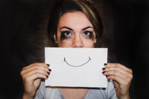 Woman with pale skin and dark shadows beneath her eyes holding up a drawing of a smile in front of her mouth.