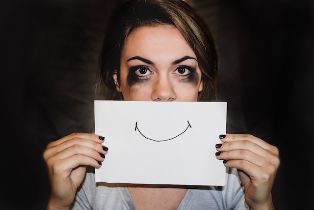 Woman with pale skin and dark shadows beneath her eyes holding up a drawing of a smile in front of her mouth.