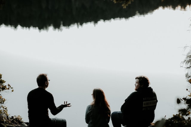 Silhouette of three people sitting on a cliff, in front of some water.
