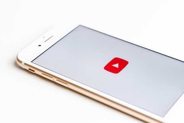 A rose gold iPhone against a white background. The screen shows a red YouTube play button.
