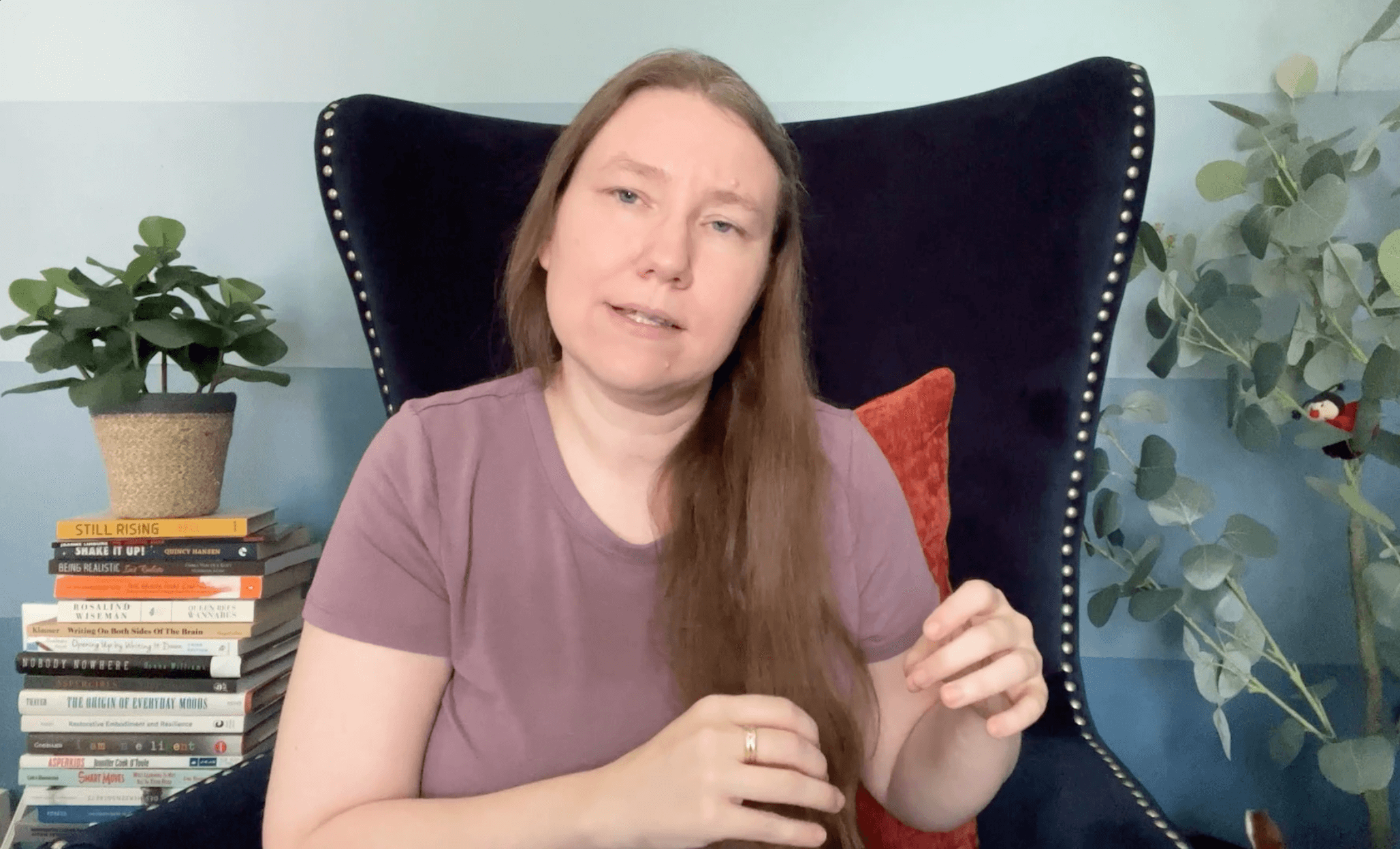 Picture of a pale skinned woman in a purple shirt with long hair, talking at the camera and gesturing with her hands. Next to her is a large plant and a pile of books and a blue wall behind her.
