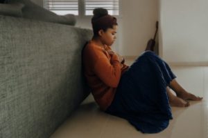 Woman with dark skin and hair pulled up in a bun, wearing an orange shirt and black skirt, sits on the floor against a couch with her knees pulled up and arms crossed, looking anxious. In the background a guitar leans against a wall.