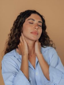 A latina woman with her hands on her neck and eyes closed, wearing a light blue shirt and a plain background.