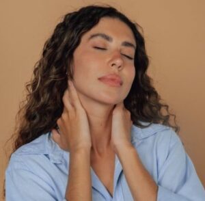 A latina woman with her hands on her neck and eyes closed, wearing a light blue shirt and a plain background.