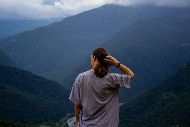 Back of a male presenting person who is scratching their head and looking at a mountain range in hazy blue colors as if he is wondering where to go.