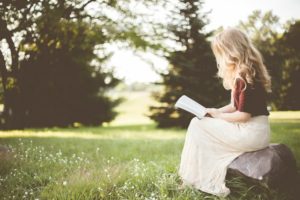 Person sitting on a rock, looking at a book by some trees.