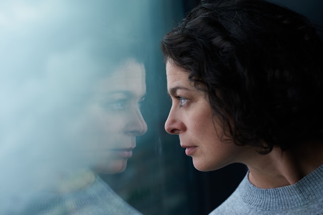 Pale skinned woman with dark hair looks anxious at her reflection in a window.