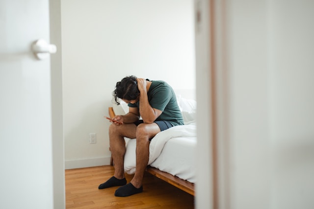 A man sits on the edge of a bed, hunched over, looking anxiously at his phone.