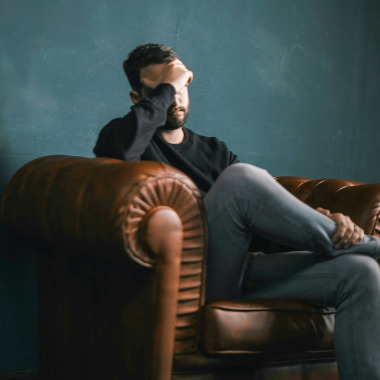A male presenting person sitting on a leather couch with his head in his hand, looking in troubled.
