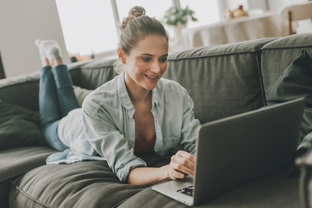 Someone laying on their stomach on a gray couch, smiling as they type on the computer in front of them.