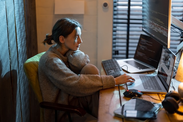 Someone sitting with their legs curled up at a desk, staring at a computer with multiple monitors in front of them.