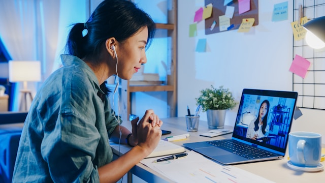 Someone sitting with their arms rested on their desk. They have earbuds in, and are looking at their computer screen that has a person talking on it.
