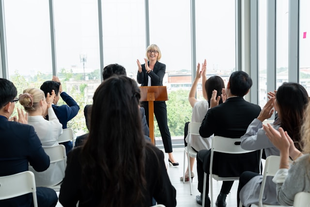 A woman standing and smiling at a podium in an office setting. There's a couple rows of people in front of her, clapping their hands.