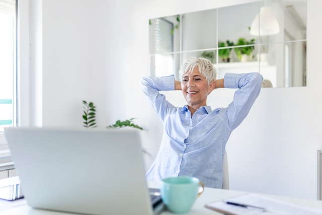 A person sitting at a desk in a room with bright window light. They are holding their hands behind their head while smiling down at their gray computer in front of them.