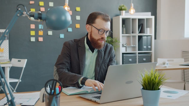 A man sitting down at a desk in an organized office, looking down and typing on a computer.