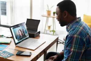 A person smiling and facing towards a laptop rested on top of a wooden table. The laptop displays a video call with another person.