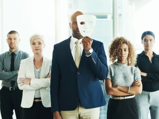 Group of people in formal attire standing in a line, staring at the camera. The person in the center is holding a white mask to their left eye.