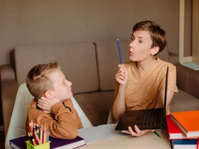 A kid and an adult at a desk. The kid is looking towards the adult, while the adult is looking at a pencil they are holding to their face, as well as holding a computer.