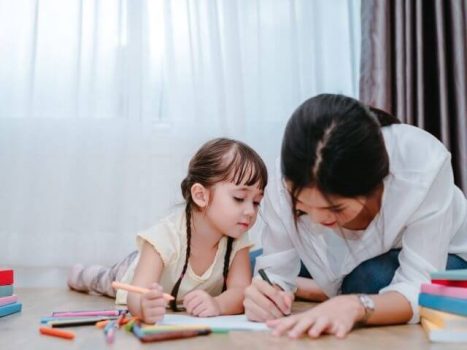 Kid and adult laying on the floor, writing on some paper. There's scattered, multicolour pencils to their sides.
