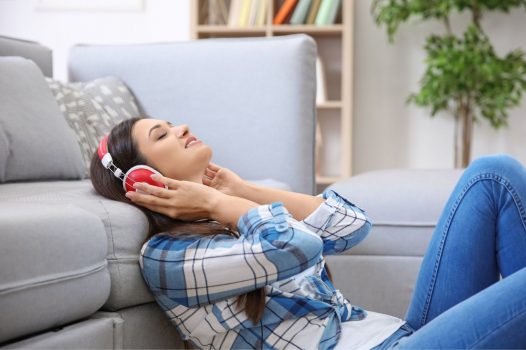 Person leaning up against a grey couch, holding their hands to their red headphones.
