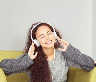 Person in a grey jumper, sitting on a lime couch, holding their hands to their headphones and smiling.