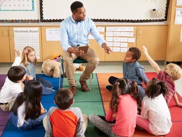 Group of kids sitting in a circle with an adult sitting on a chair at the outer middle, pointing at a kid in a red shirt with their hand raised.
